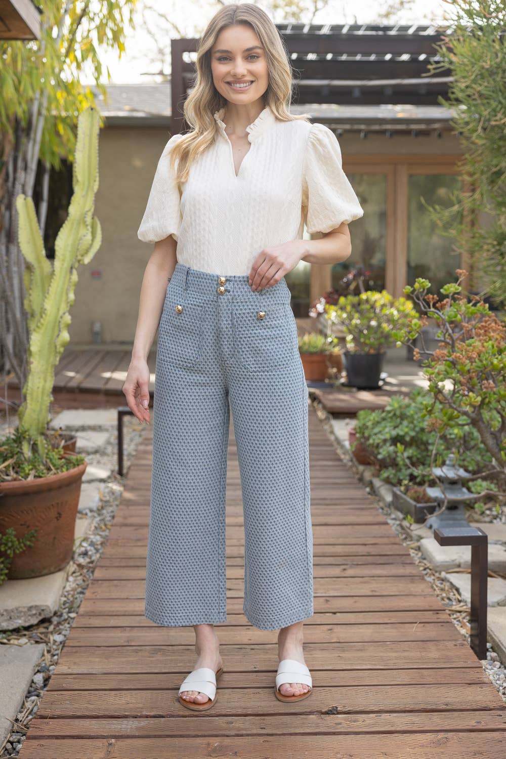 Woman in a white blouse and blue checkered pants standing on a wooden path in a garden.