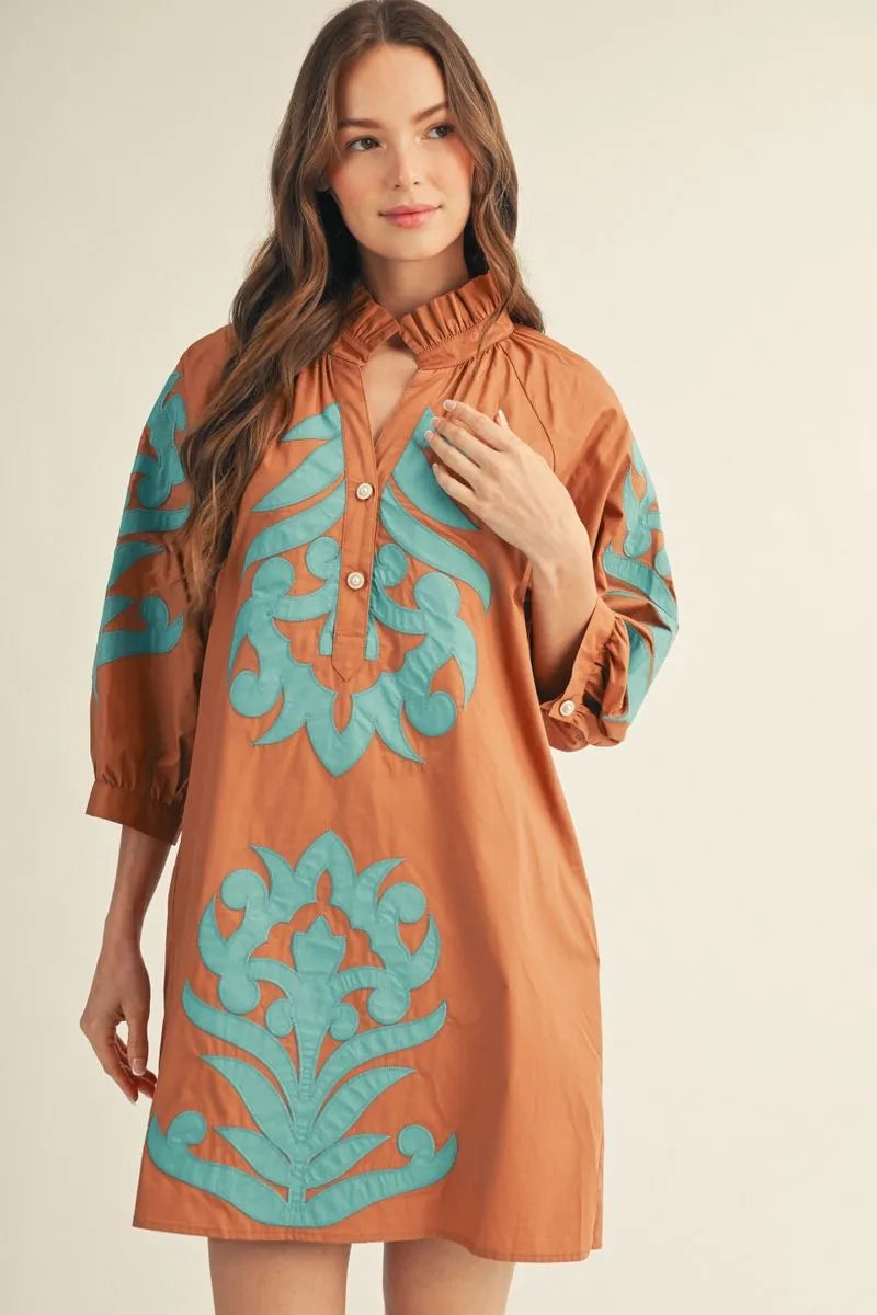 Woman wearing a rust cotton shift dress with teal baroque floral appliqué detail, ruffled high neckline, and 3/4 sleeves — Mirror on Main