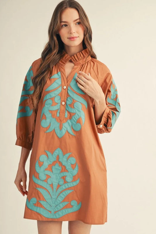 Woman wearing a rust cotton shift dress with teal baroque floral appliqué detail, ruffled high neckline, and 3/4 sleeves — Mirror on Main