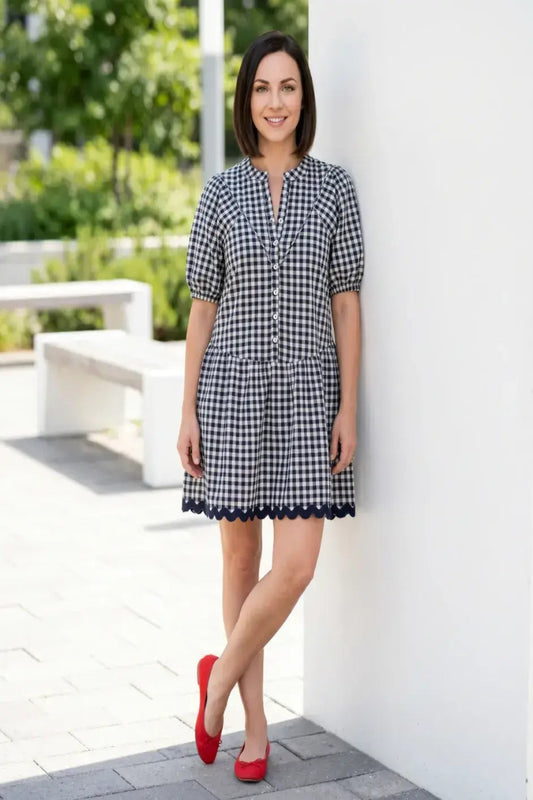 Woman wearing a navy and white gingham cotton mini dress with a mandarin neckline, shell button front, short puff sleeves, ric rac trim, and above-the-knee hem.