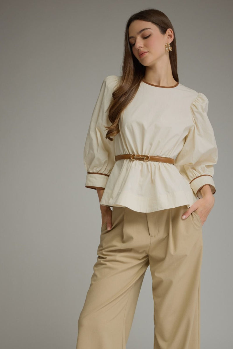 A woman modeling a cream puff-sleeve blouse with a tan belt and beige wide-leg trousers, standing against a neutral gray studio background.