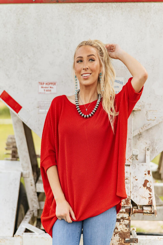 Woman wearing a red sweater and blue jeans standing in front of a rustic background.