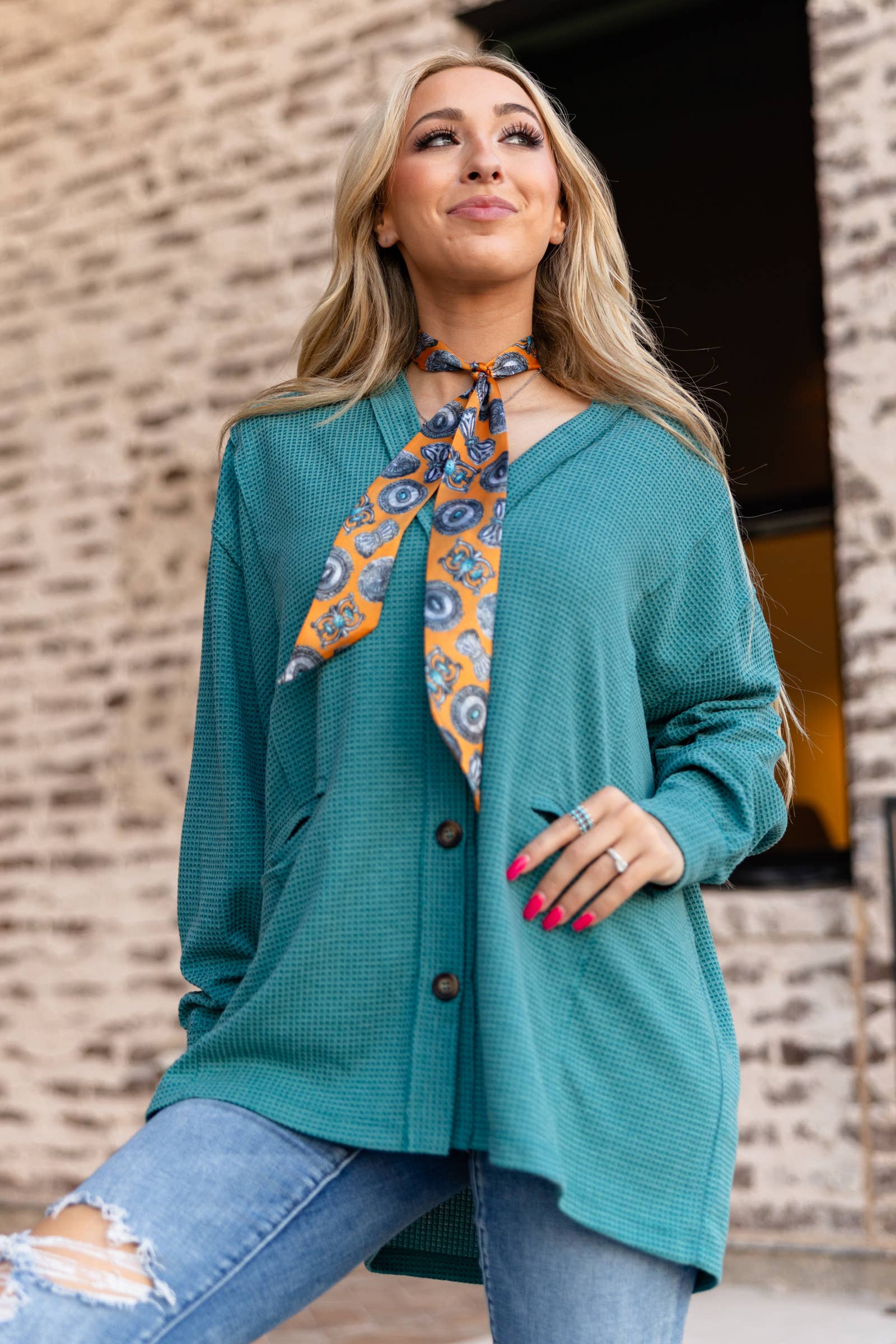 Woman wearing a teal cardigan with a patterned scarf, standing against a brick wall.