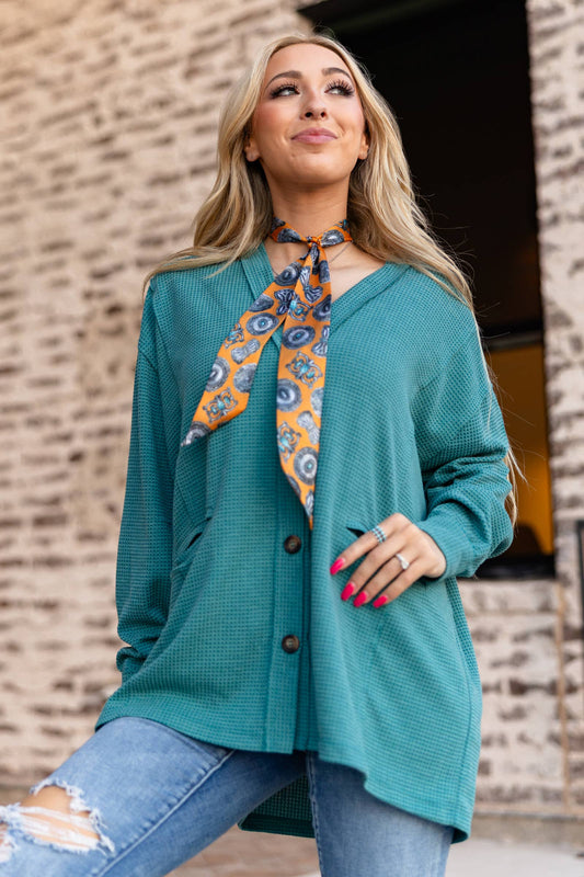Woman wearing a teal cardigan with a patterned scarf, standing against a brick wall.