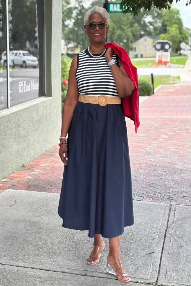Woman wearing a striped sleeveless top and navy skirt holding a red jacket at Mirror On Main.
