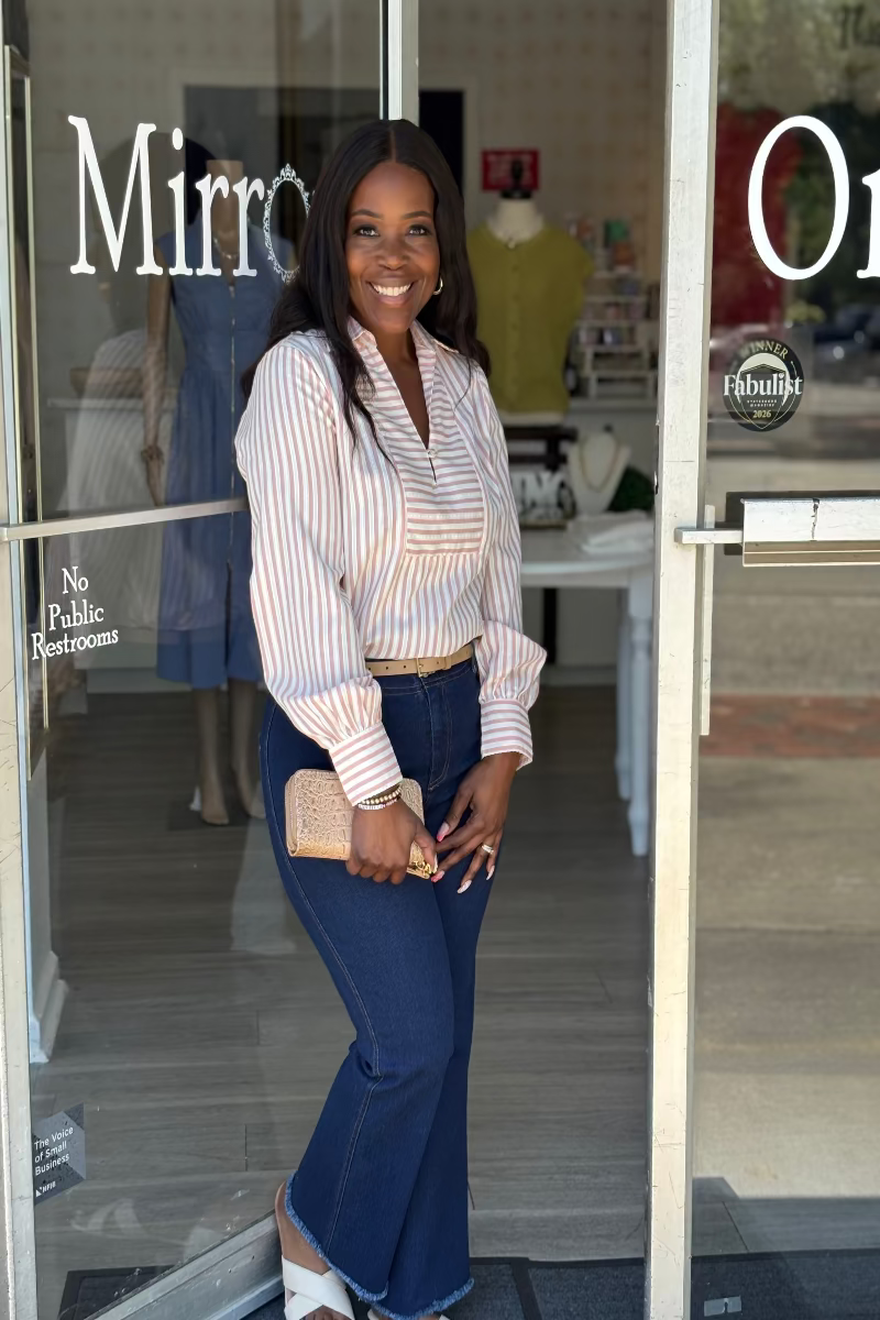 Woman standing outside a store with glass doors, wearing a striped shirt and blue jeans. Mirror On Main