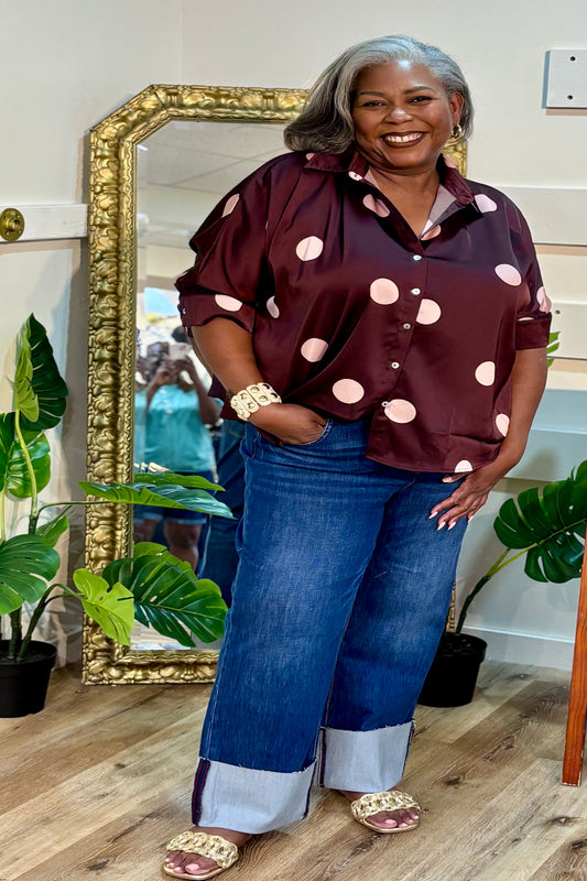 Plus size woman wearing a plum and blush pink polka dot collared button-front top with 3/4 sleeves, styled with dark wide-leg cuff jeans and flat gold shoes— Mirror on Main