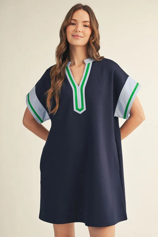 Woman wearing a navy relaxed shift dress with a split V-neckline featuring white and green contrast trim, drop shoulders, short sleeves with contrast cuffs, and side pockets.