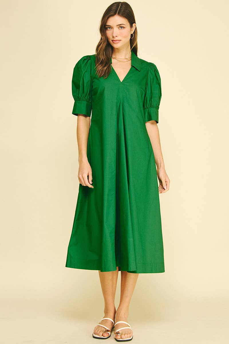 Woman wearing an emerald green midi dress with a collared V-neckline, pleated front detail, short balloon sleeves, wide cuffs, relaxed silhouette, and side pockets.