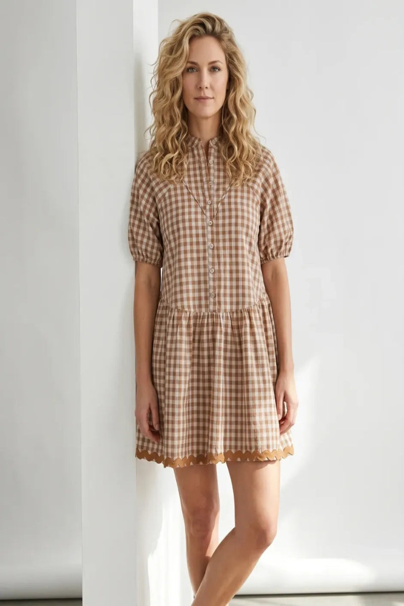 A woman with blonde curly hair wearing a brown and beige gingham mini dress with puff sleeves, standing against a white wall.