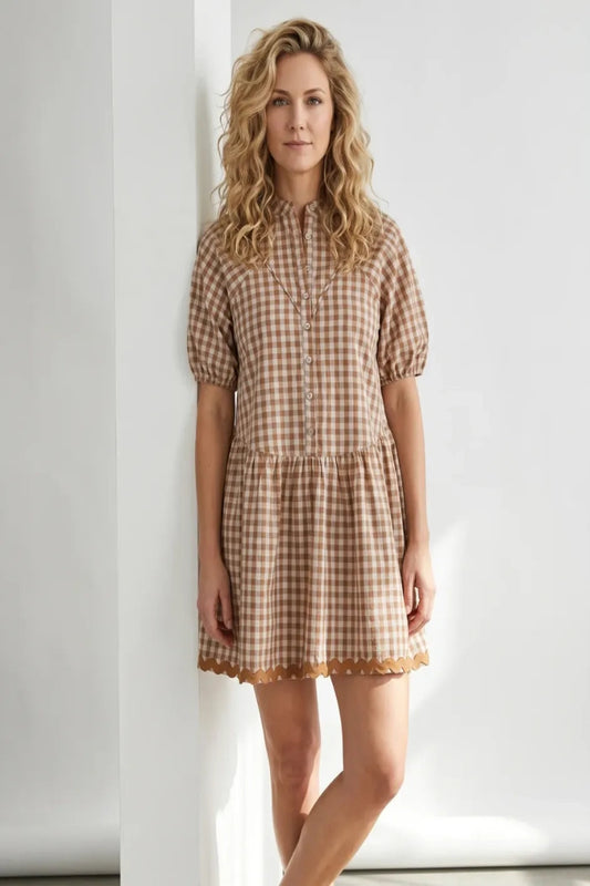 A woman with blonde curly hair wearing a brown and beige gingham mini dress with puff sleeves, standing against a white wall.